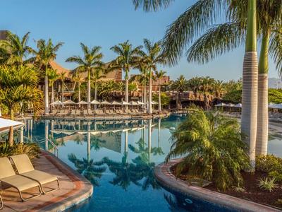 Klidná oblast u bazénu s palmami, lehátky a jasnou odrazovou vodou pod modrou oblohou.