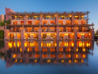 Modernes Gebäude mit Balkonen, Beleuchtung und Reflexion in einem ruhigen Wasserbecken bei Sonnenuntergang.