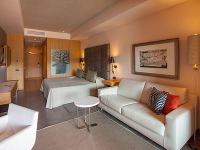 Modern eingerichtetes Hotelzimmer mit Doppelbett, Sofa, zwei Tischen und warmen Lichtquellen.