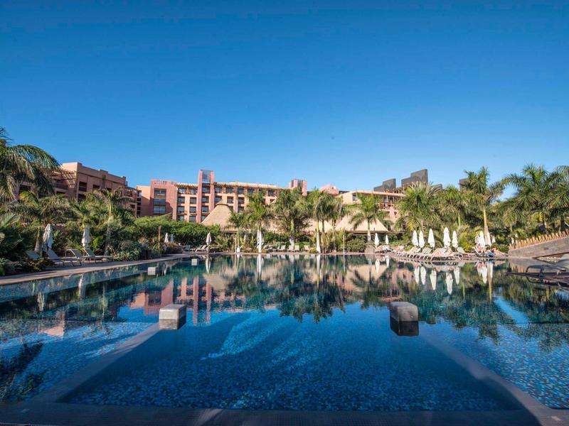 Niebieski basen odbijający palmy i hotel pod jasnym niebem.