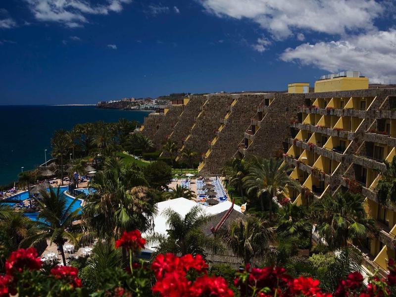 Complesso alberghiero con edifici a terrazza, palme e piscina vicino al mare sotto un cielo blu.