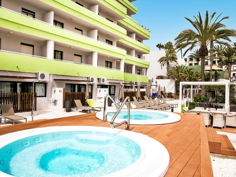 Buitenspa's bij een hotel met houten terras en palmbomen onder een blauwe lucht.