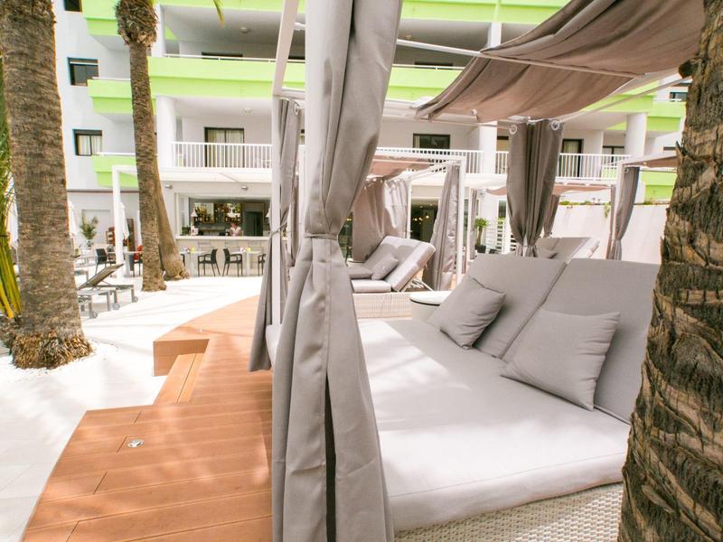 Buitenloungebed met kussens onder luifel bij palmbomen op hotelterras.