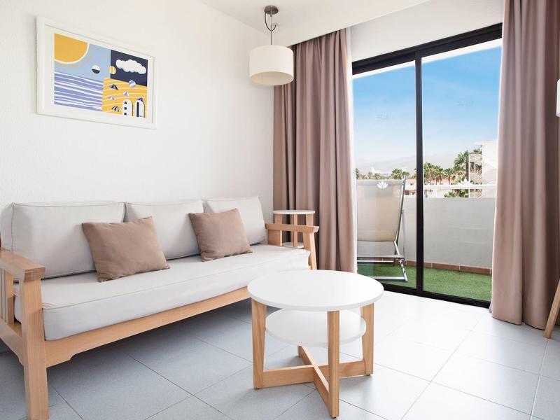 Lichte woonkamer met meubels van licht hout, beige kussens, witte ronde tafels en glazen deuren naar balkon.