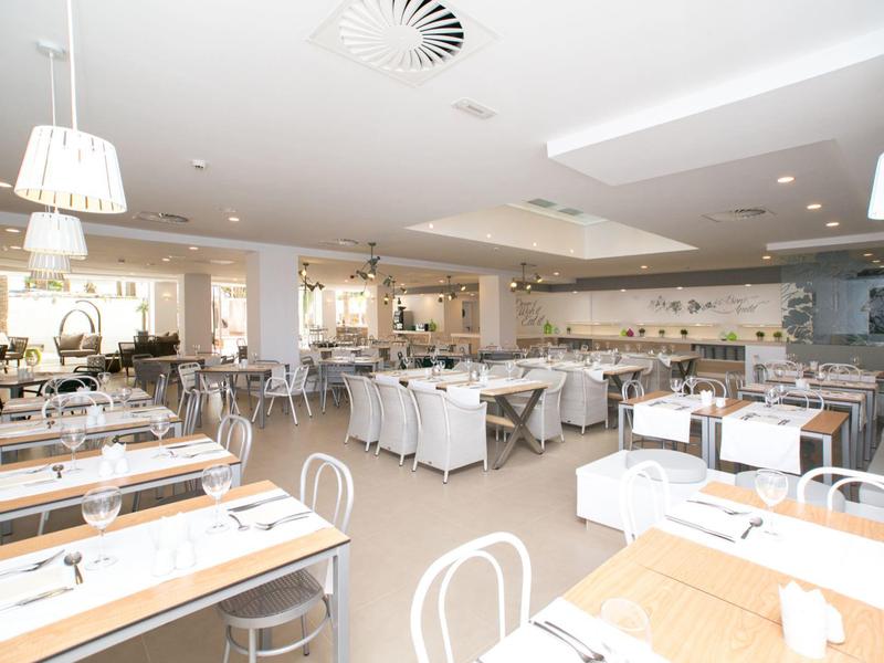 Lichte, ruime hotelrestaurant met modern wit meubilair, grote ramen en hanglampen.