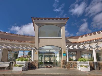 Ingresso dell'hotel con facciata in vetro, pergola e cielo ampio di giorno.