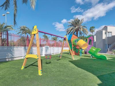 Parco giochi per bambini con altalene e scivolo su prato artificiale verde sotto un cielo limpido