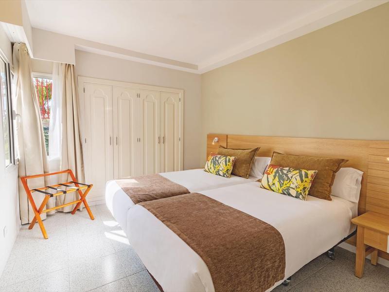 Camera d'hotel luminosa con due letti singoli, testiera in legno e piccola sedia vicino alla finestra.