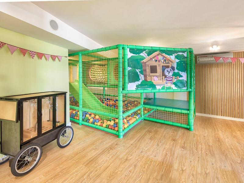 Area giochi interna colorata con struttura per arrampicata e carrello di legno su pavimento in legno.