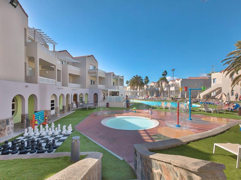 Hotel moderno con piscina all'aperto, parco giochi e scacchi sul prato sotto un cielo limpido.