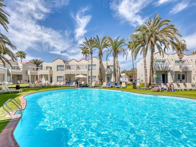 Piscina all'aperto luminosa con palme e edifici dell'hotel a due piani sotto un cielo azzurro.
