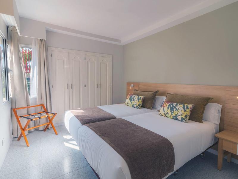 Camera d'hotel luminosa con letto matrimoniale, comodini, sedia e armadio in tonalità pastello.