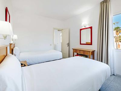 Chambre lumineuse avec deux lits simples, linge blanc, miroir et fenêtre avec vue sur des palmiers.