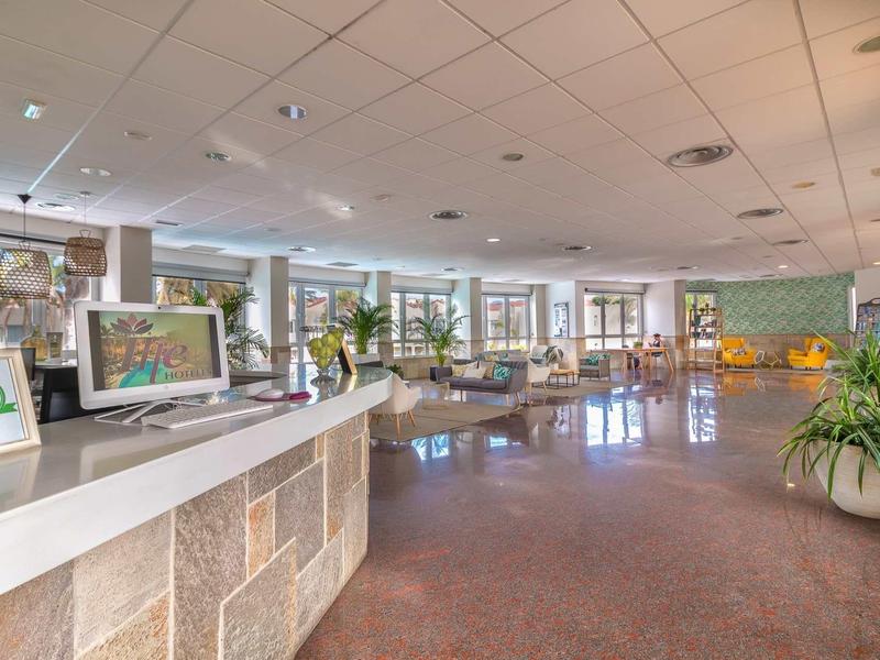 Hall d'hôtel lumineux avec comptoir d'accueil, plantes et coin salon.