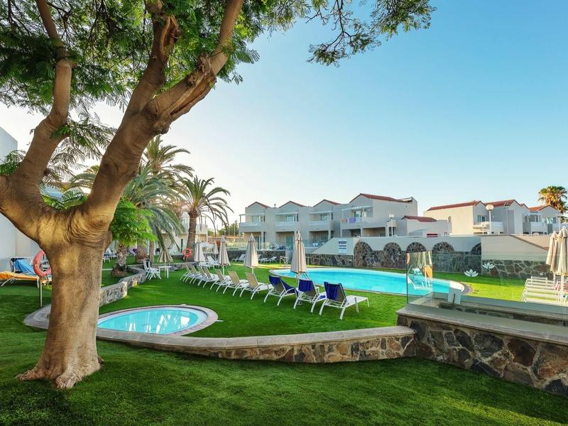 Jardin verdoyant avec piscine, chaises longues et bâtiments blancs sous un ciel bleu clair.