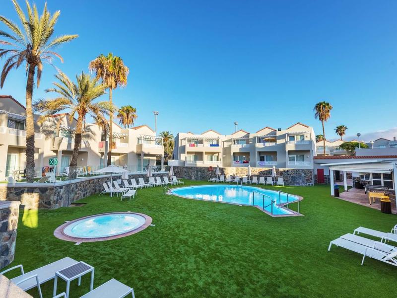 Piscine d'hôtel avec terrasse ensoleillée, chaises longues et palmiers sous un ciel bleu clair