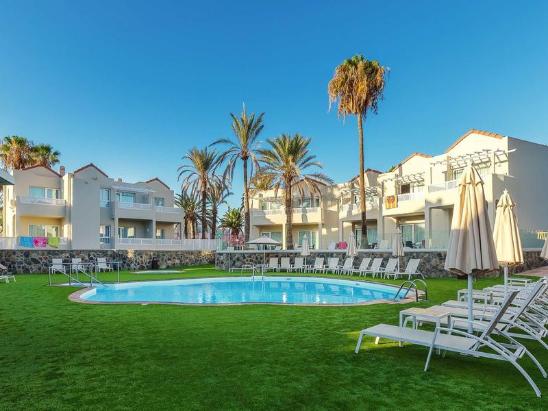 Complexe hôtelier avec piscine, chaises longues et parasols sur pelouse verte sous ciel clair.