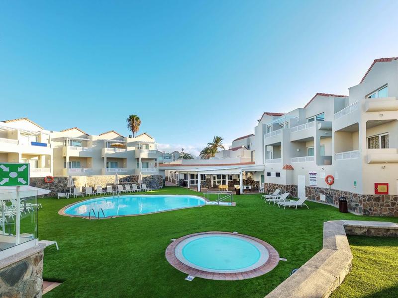 Complexe hôtelier avec deux piscines, bâtiments blancs et pelouse verte sous un ciel bleu.