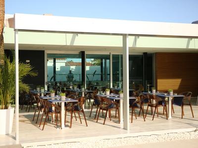 Freiluft-Café mit weißen Stühlen, Tischen und großen Glasfronten unter blauem Himmel.