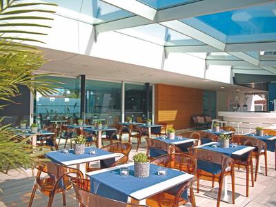 Freiluftrestaurant mit blauen Tischdecken, Holztischen und Stühlen unter Glasdach und Pflanzen.