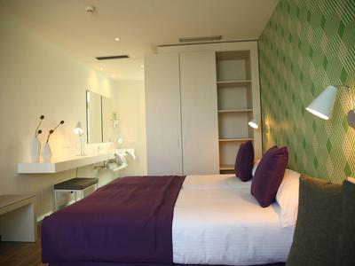 Modernes Schlafzimmer mit großem Bett, lila Bettdecke, grüner gemusterter Wand und elegantem Waschbereich.