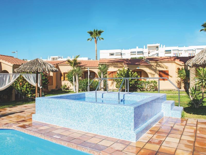 Moderner Außenpool mit klarem Wasser, Sonnenschirmen und Palmen vor Gebäuden unter blauem Himmel.