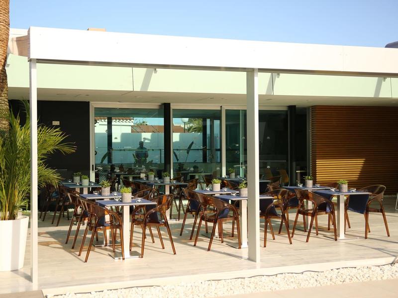 Freiluft-Café mit weißen Stühlen, Tischen und großen Glasfronten unter blauem Himmel.
