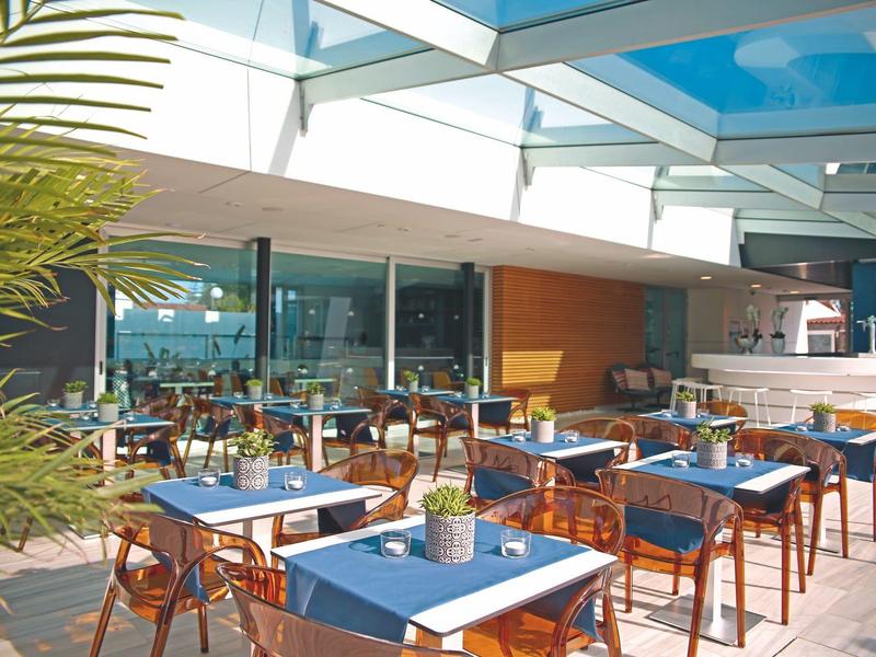 Freiluftrestaurant mit blauen Tischdecken, Holztischen und Stühlen unter Glasdach und Pflanzen.