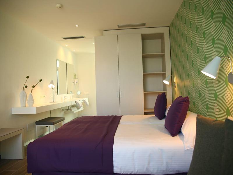 Modernes Schlafzimmer mit großem Bett, lila Bettdecke, grüner gemusterter Wand und elegantem Waschbereich.