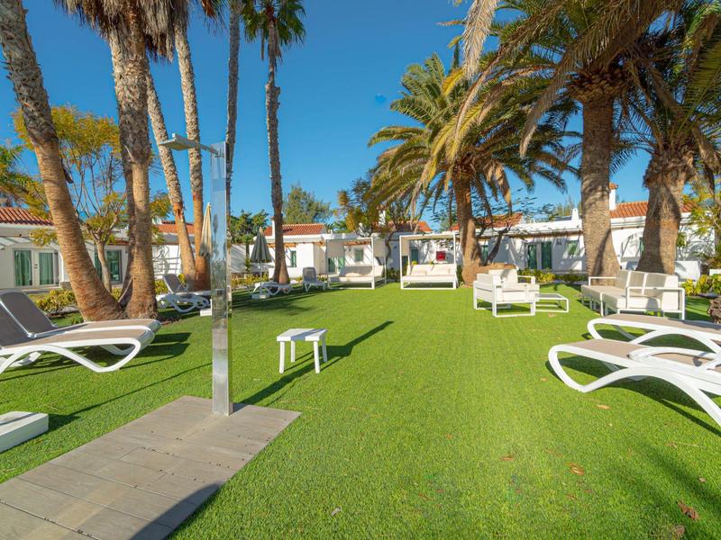 Prato verde con lettini e palme davanti a bungalow bianchi sotto un cielo azzurro.