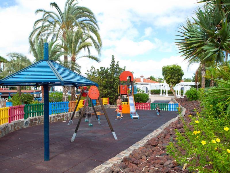 Parco giochi colorato con altalene, scivoli e ombrellone in un giardino soleggiato.