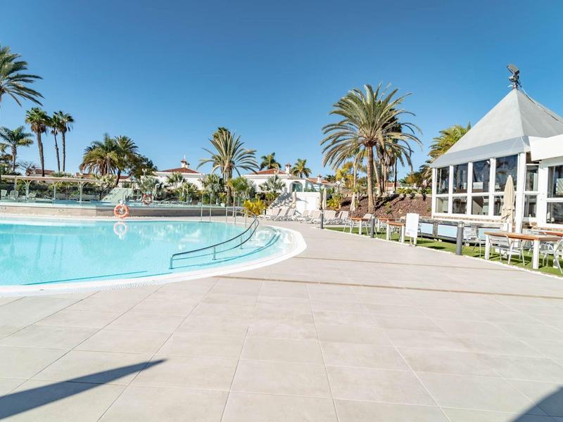 Grande piscina all'aperto con terrazza soleggiata e palme sotto un cielo azzurro.