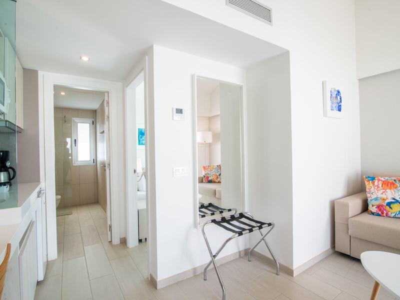 Camera d'hotel luminosa con porta valigie, divano con cuscini colorati e vista su corridoio e bagno.