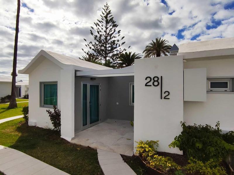 Biały dom z płaskim dachem, zielonymi drzwiami i zadbanym ogrodem pod pochmurnym niebem.