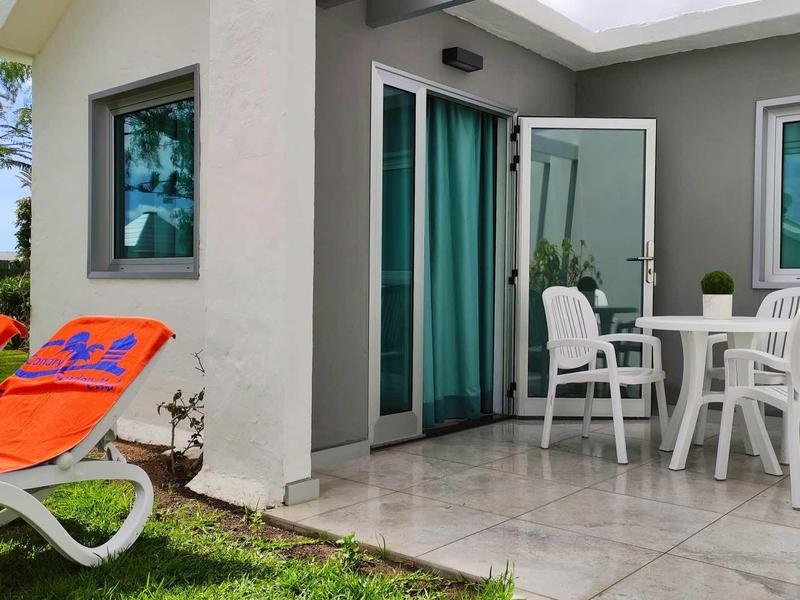 Nowoczesny pokój hotelowy z tarasem, białymi meblami i pomarańczowymi leżakami na trawniku.