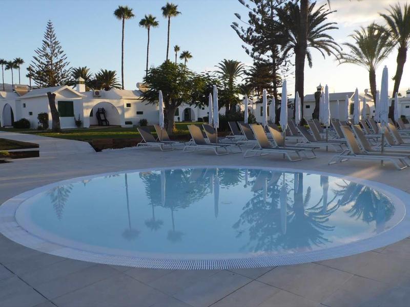 Okrągły basen otoczony białymi leżakami i palmami w hotelowym ogrodzie o zachodzie słońca.