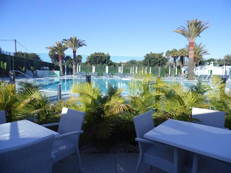 Nowoczesny teren zewnętrzny z stołami, krzesłami, palmami i basenem pod czystym niebieskim niebem.