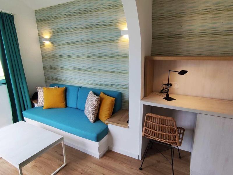 Jasny pokój hotelowy z niebieską sofą, kolorowymi poduszkami, biurkiem i krzesłem obok okna z zasłoną.