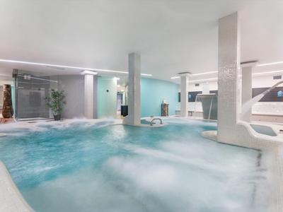 Großes Hallenbad mit Whirlpool, Wasserfall und modernen Säulen in hellen Blautönen.