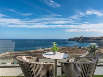 Balkon mit zwei Stühlen, Tisch und Blick auf das Meer unter blauem Himmel.