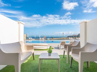 Balkon mit weißem Mobiliar, grünem Kunstgras und Blick auf Pool und Meer unter blauem Himmel.