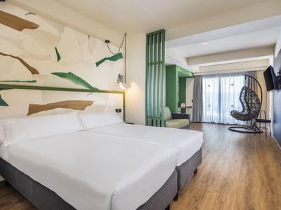 Modernes Hotelzimmer mit zwei weißen Betten, Holzboden, grünem Akzent und einem hängenden Schaukelstuhl.