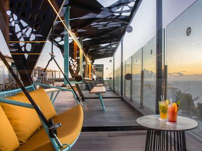 Moderní střešní terasa s barevnými křesly a výhledem na město a moře při západu slunce.