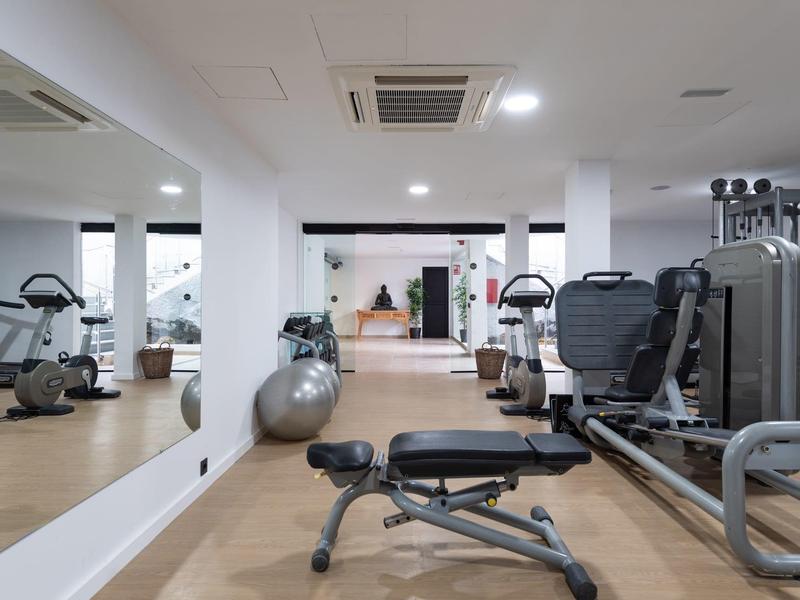 Moderner Fitnessraum mit Hantelbänken, Trainingsgeräten, großer Spiegelwand und hellem Holzboden.