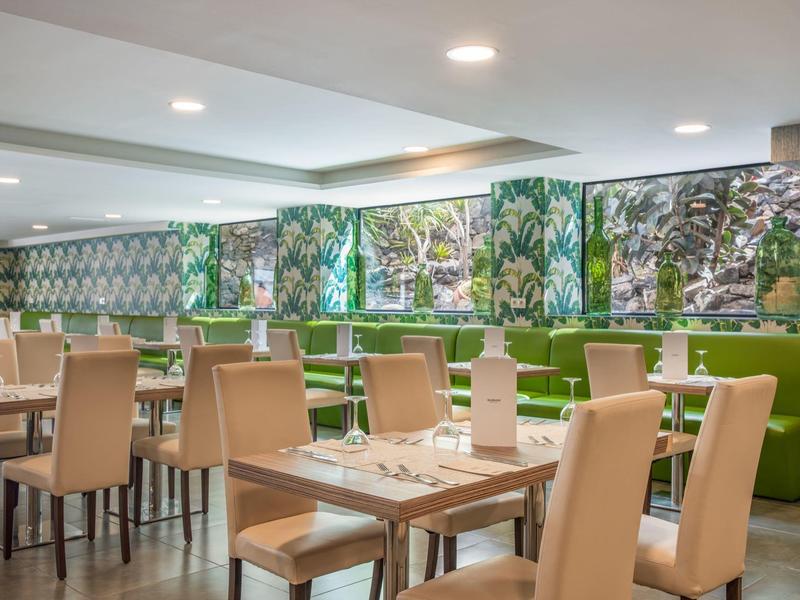 Modern eingerichteter Speisesaal mit hellen Stühlen, Holztischen und großem Pflanzenmotiv an der Wand.