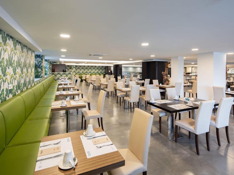 Großer, moderner Speisesaal mit weißen Stühlen, Holztischen und grün gemusterter Wand rechts.