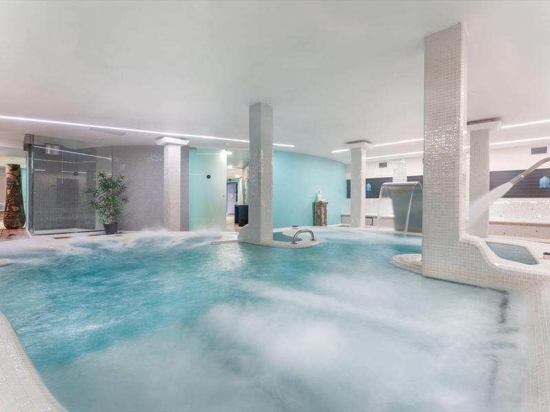 Großes Hallenbad mit Whirlpool, Wasserfall und modernen Säulen in hellen Blautönen.