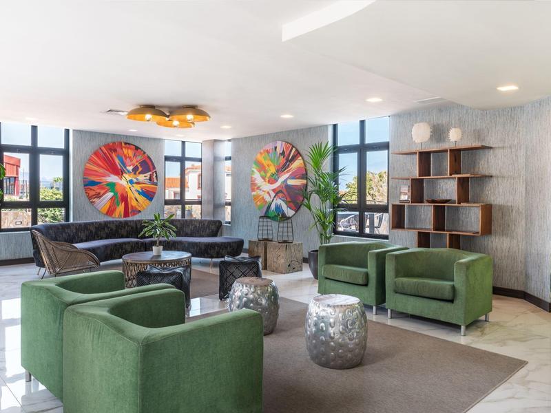 Modernes Wohnzimmer mit grünen Sesseln, schwarzem Sofa, bunten Wandbildern und großem Fenster.