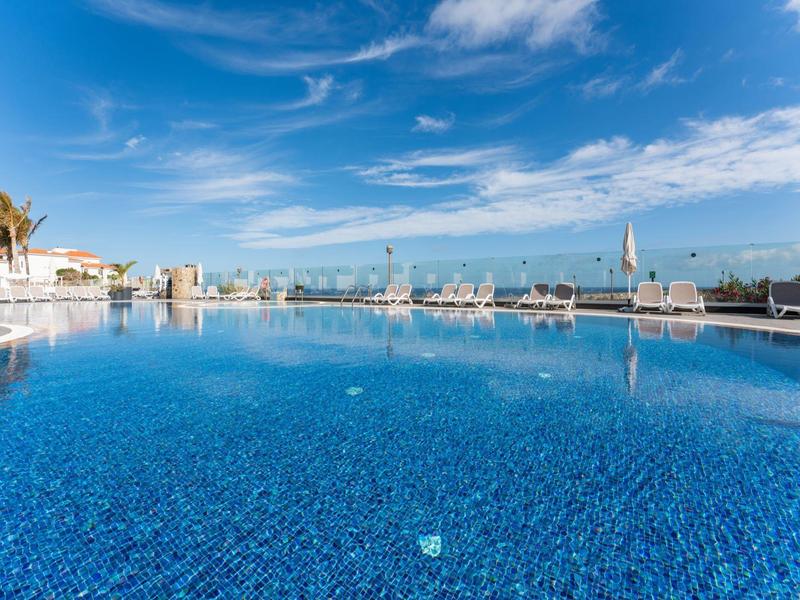 Großer blauer Außenpool mit klarem Wasser, Sonnenliegen und palmbaumunter blauem Himmel.