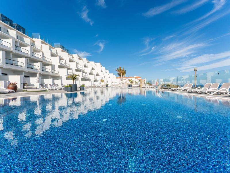 Großer blauer Pool mit weißen Liegestühlen und modernem, weißem Hotelgebäude unter blauem Himmel.